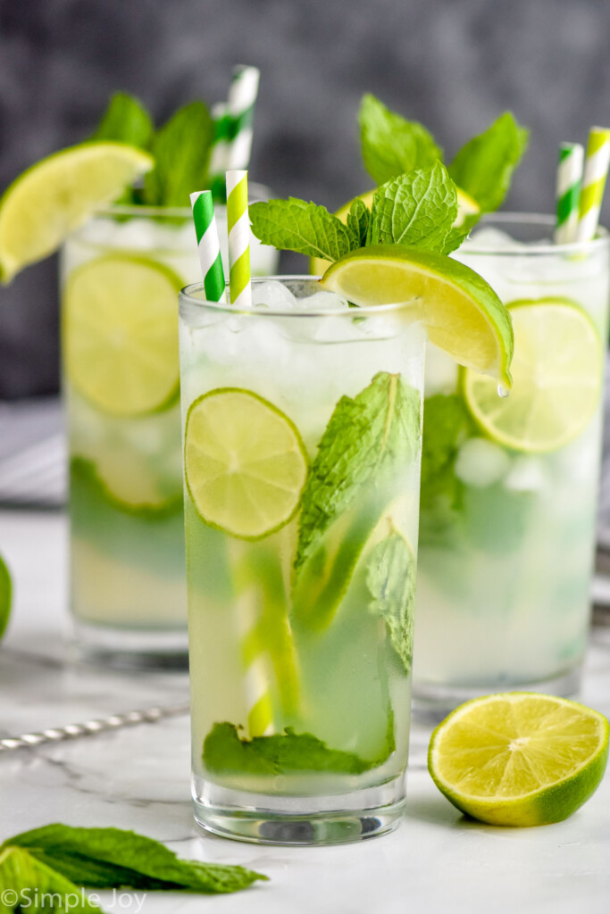 three high ball glasses on a white surface against a gray background filled with lime slices, a mojito recipe, fresh mint leaves, and garnished with green and white stripped straws, fresh mint and a lime wedge