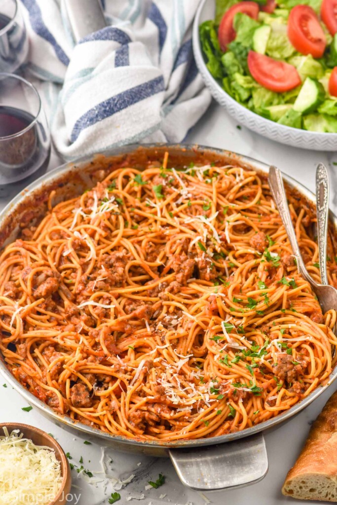 Large skillet of Sausage Spaghetti garnished with parsley and shredded parmesan cheese with salad and glasses of wine beside