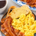 Pinterest graphic for Bacon in The Oven. Text says "how to make bacon in the oven simplejoy.com" Image shows plate of bacon cooked in the oven, scrambled eggs, and toast. Plate of bacon, mug of coffee, and fresh berries surrounding.