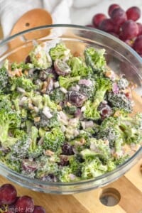 Overhead view of Broccoli Salad in a glass bowl with wooden spoon and grapes beside.