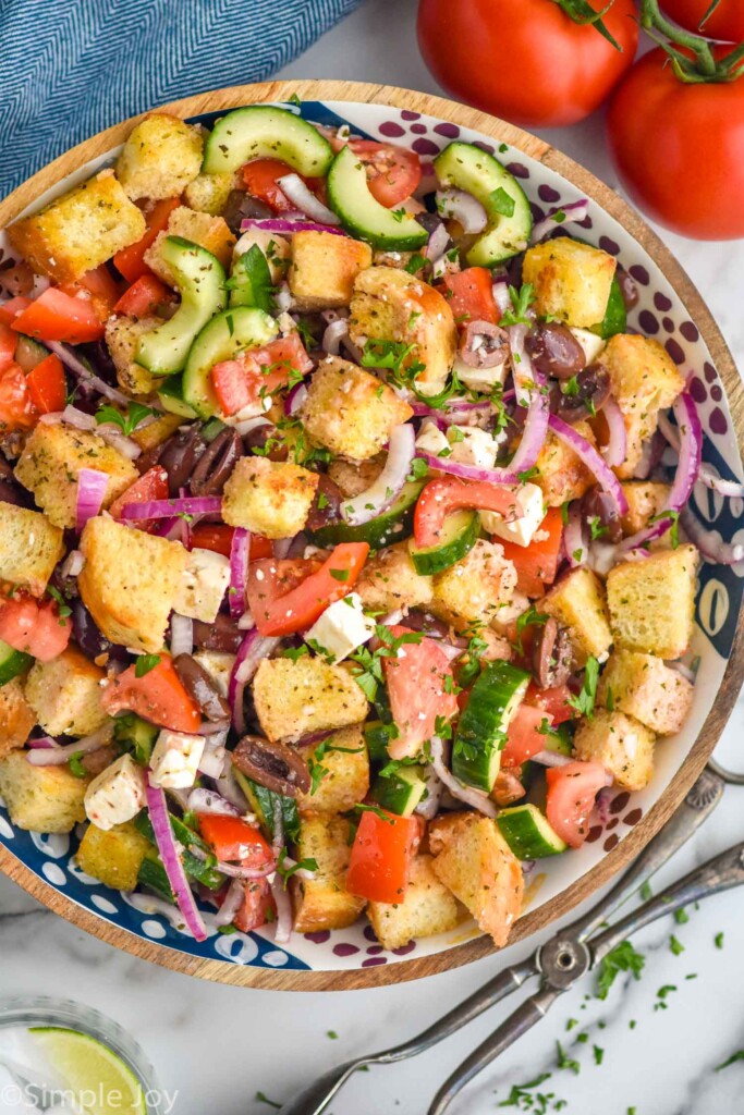 overhead of bowl of Greek Panzanella Salad with tomatoes and serving tongs sitting beside