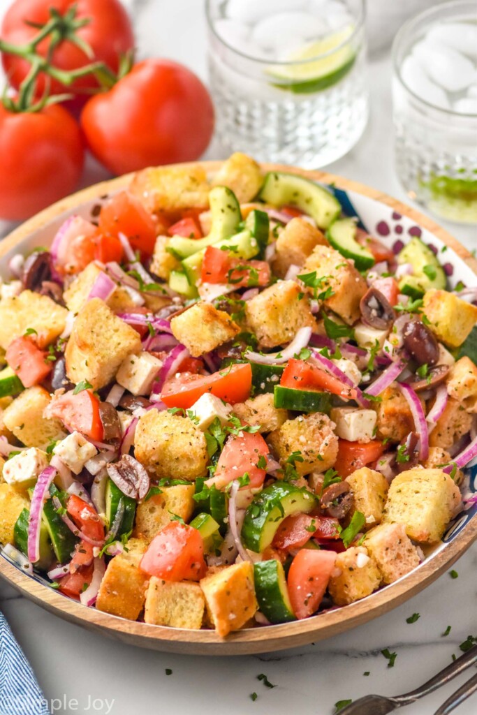 bowl of Greek Panzanella Salad with tomatoes and two drinking glasses sitting in background