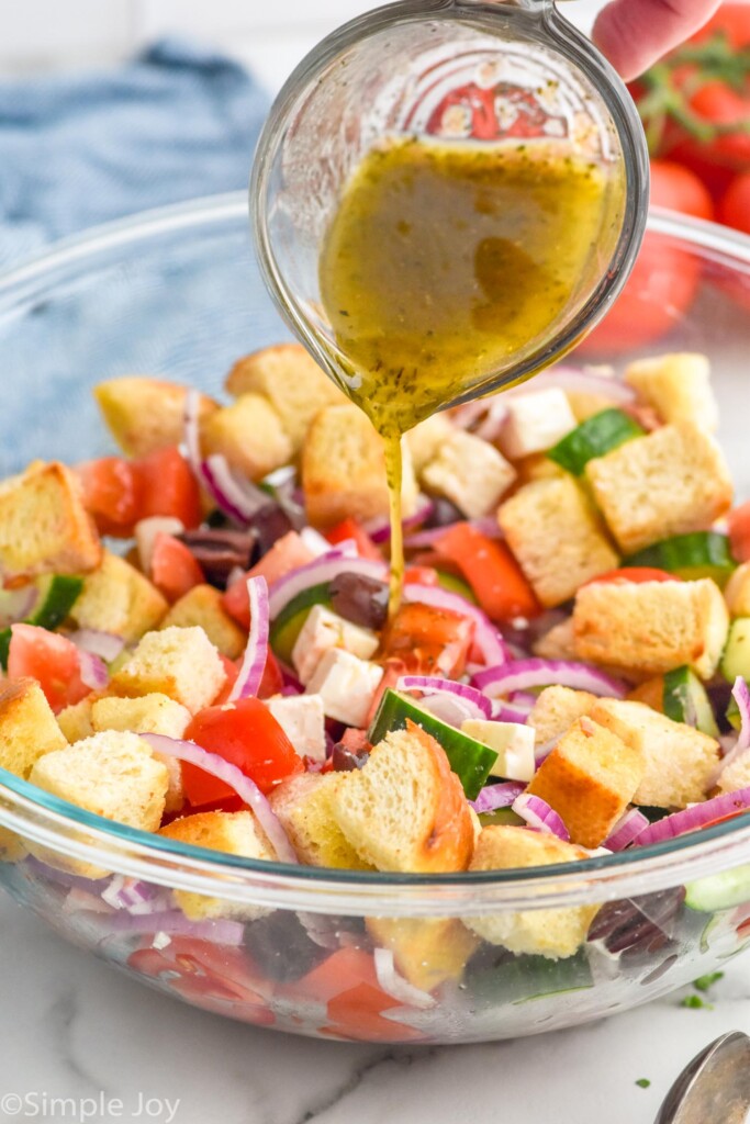 glass measuring cup pouring dressing over bowl of Greek Panzanella Salad