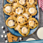 Pinterest graphic for S'mores Cookies recipe. Image shows a platter of S'mores Cookies with glasses of milk, chocolate chips, marshmallows, and graham crackers beside. Text says, "S'mores Cookies simplejoy.com"