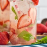 Pinterest graphic for strawberry mojito. Image shows glass of Strawberry Mojito with ice, fresh strawberry and mint garnish, and a straw. Fresh strawberries and Strawberry Mojito sitting in background. Text says "strawberry mojito simplejoy.com"