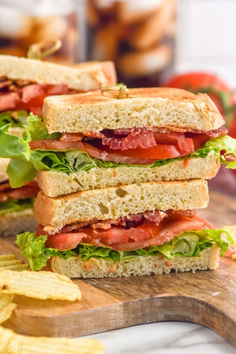 Side view of BLT sandwich cut in half and stacked on top of each other on a wooden board. Chips beside.