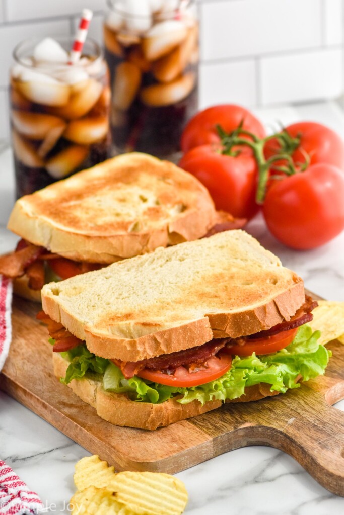 two BLT sandwiches on a wooden board with drinks, tomatoes, and chips beside.