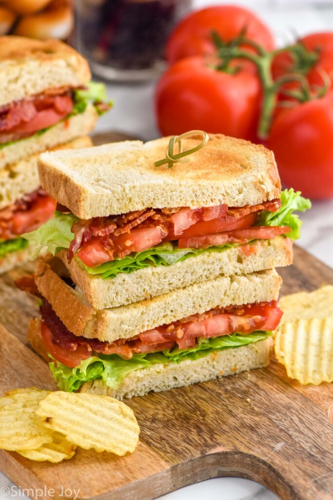 BLT sandwich cut in half and stacked on top of each other with a sandwich pick in it. Chips and tomatoes beside.