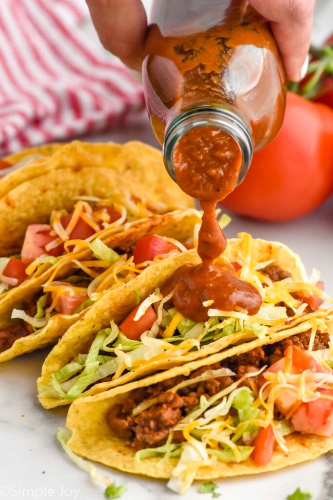 Women's hand pouring bottle of homemade taco sauce over taco shells filled with taco meat, shredded lettuce, shredded cheese, and diced tomatoes.