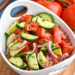 Bowl of Cucumber Tomato Salad sitting on a cutting board. Fresh tomatoes sitting in background