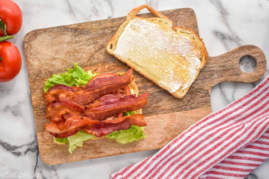 Overhead view of a BLT being constructed on a wooden board. Tomatoes and towel beside.