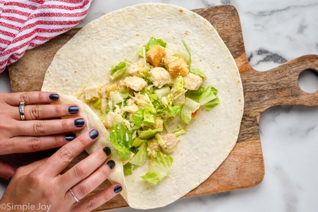Overhead view of person's hands folding side of Chicken Caesar Wrap to roll it on a wooden board