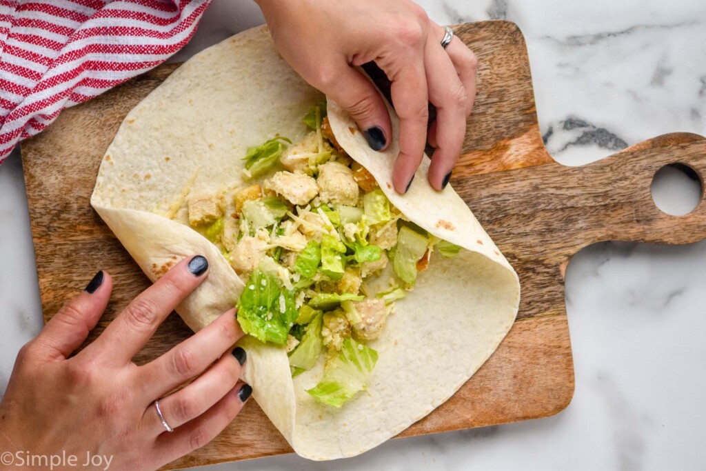 Overhead view of person's hand starting to fold a Chicken Caesar Wrap to roll it on a wooden board