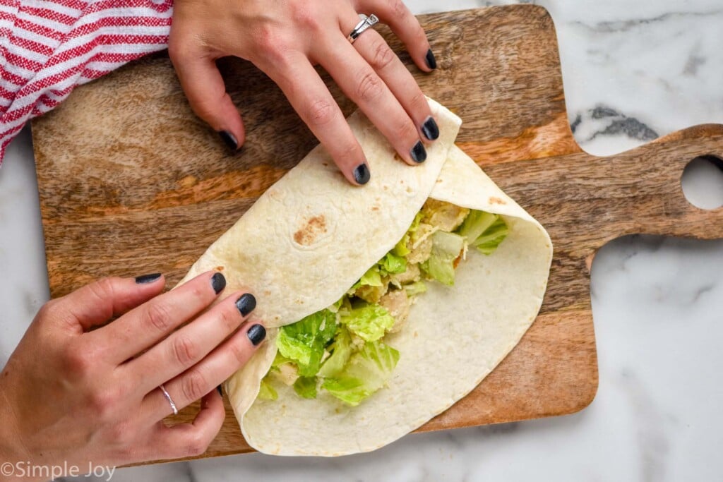 Overhead view of person's hand starting to roll Chicken Caesar Wrap on a wooden board
