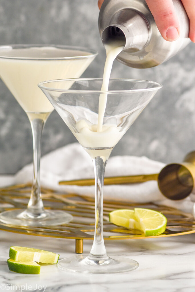 Person's hand pouring cocktail shaker of Key Lime Martini recipe into martini glass. Lime slices and cocktail jigger beside.