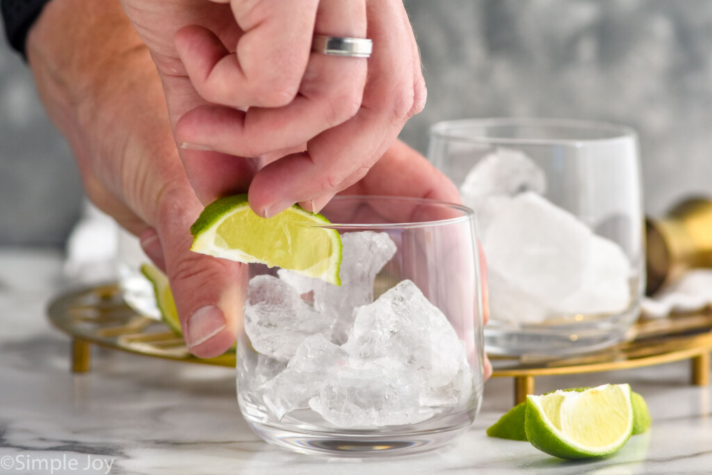 Side view of person's hand putting lime wedge on the rim of a glass with ice for Vodka Tonic recipe. Other glasses with ice and lime wedges beside.