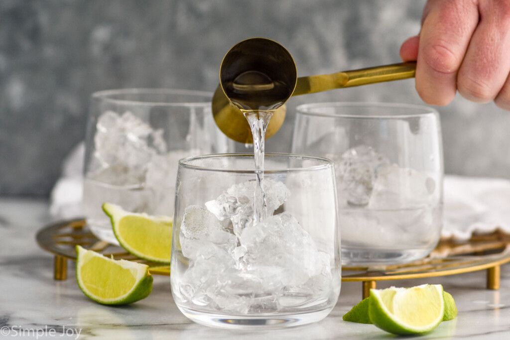 Side view of person's hand pouring cocktail jigger of vodka into glass of ice for Vodka Tonic recipe. Lime wedges beside for garnish.