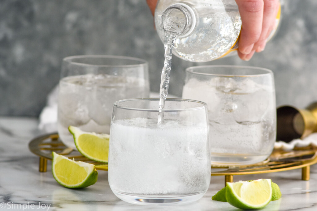 Side view of person's hand pouring tonic water into a glass for Vodka Tonic recipe. Two other glasses and lime wedges beside.