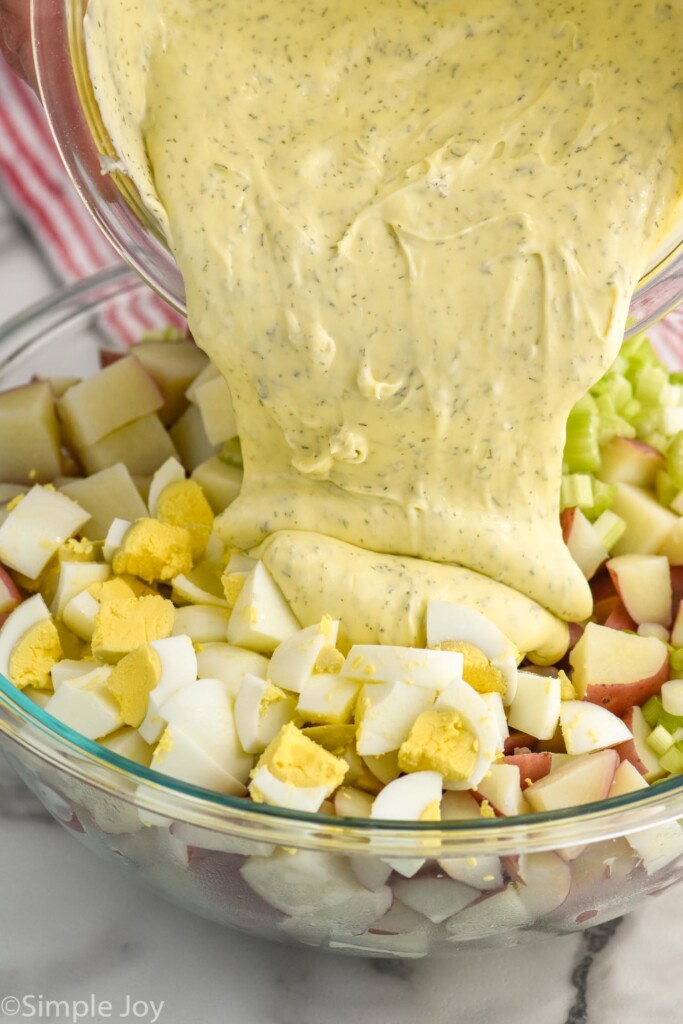 bowl of potato salad dressing pouring into bowl of potato salad ingredients.