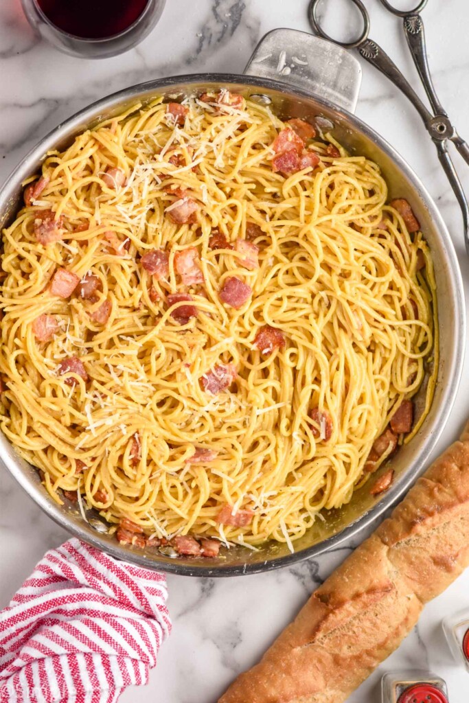 Overhead view of a large pot of Pasta Carbonara with tongs, bread, and glass of wine beside.