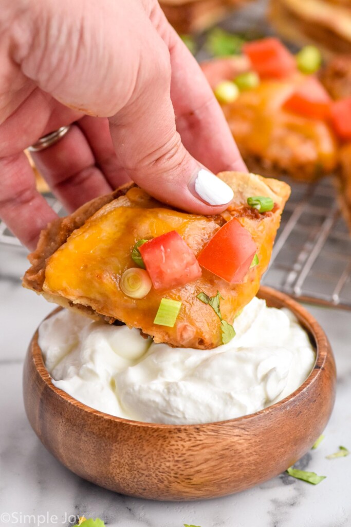woman's hand dipping piece of mexican pizza topped with diced tomato, green onions, and cilantro in a bowl of sour cream