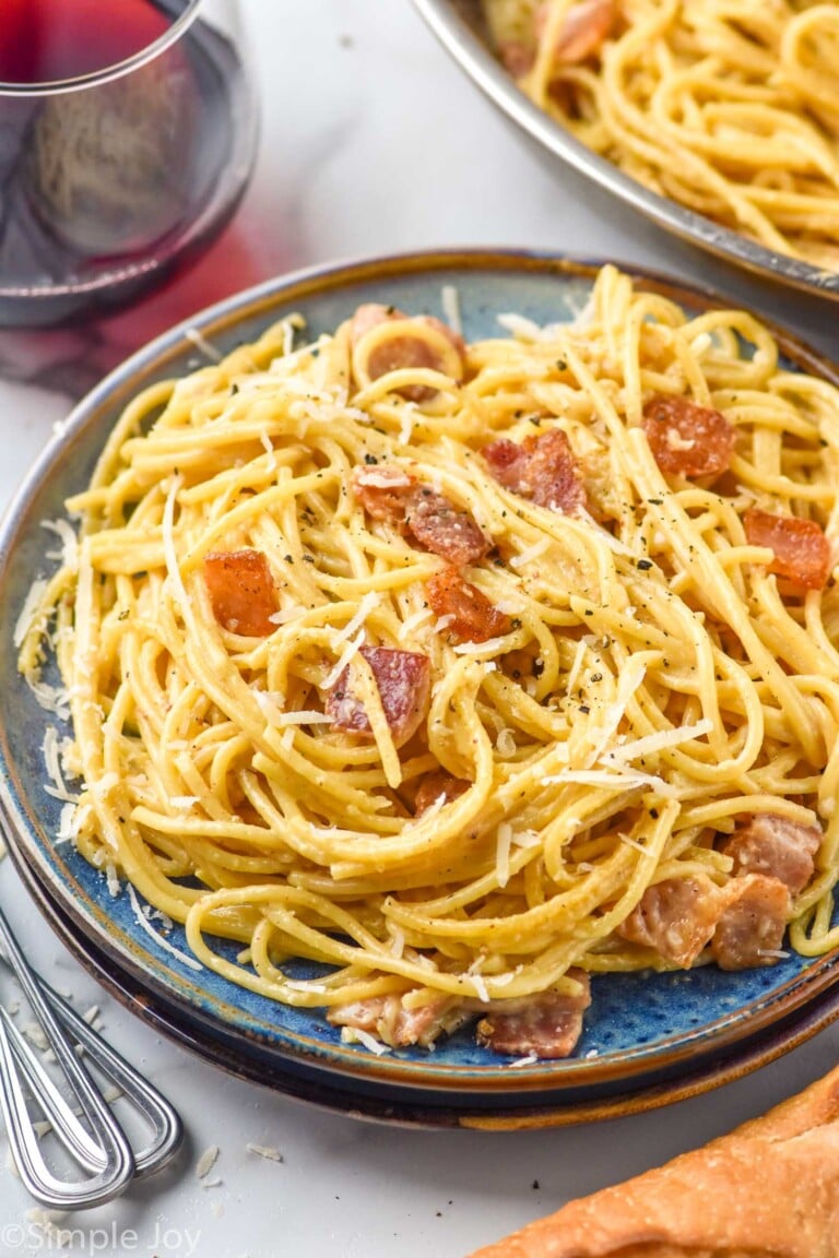 Plate of Pasta Carbonara garnished with parmesan cheese. Forks, glass of red wine, and bread beside.