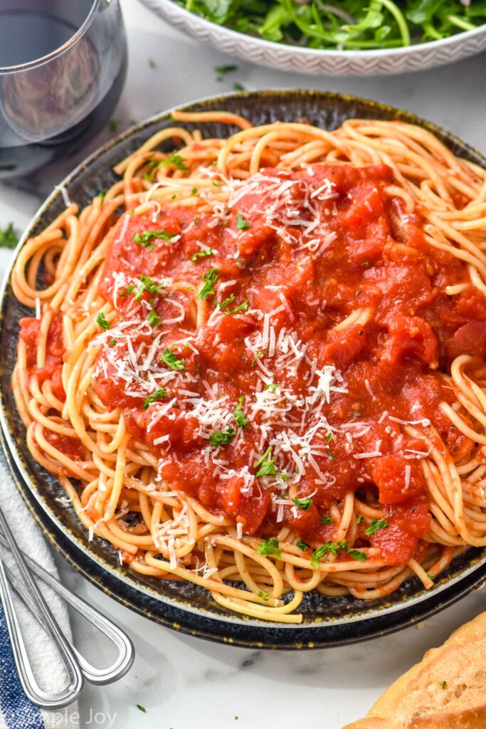 Plate of spaghetti with Spaghetti Sauce, parmesan cheese, and basil on top. Forks, glass of red wine, bread, and salad beside.
