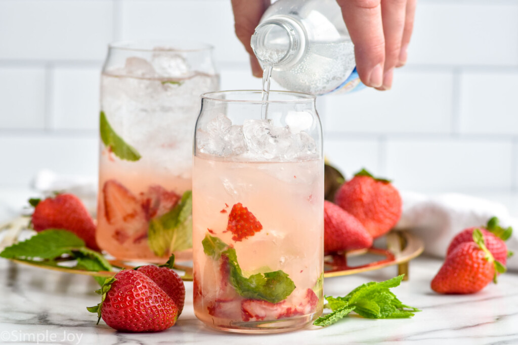 man's hand pouring club soda into a glass of strawberry mojito ingredients. Glass of strawberry mojito, fresh strawberries, and mint leaves sitting behind.