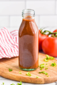 bottle of homemade Taco Sauce on a wooden board, tomatoes sitting in background