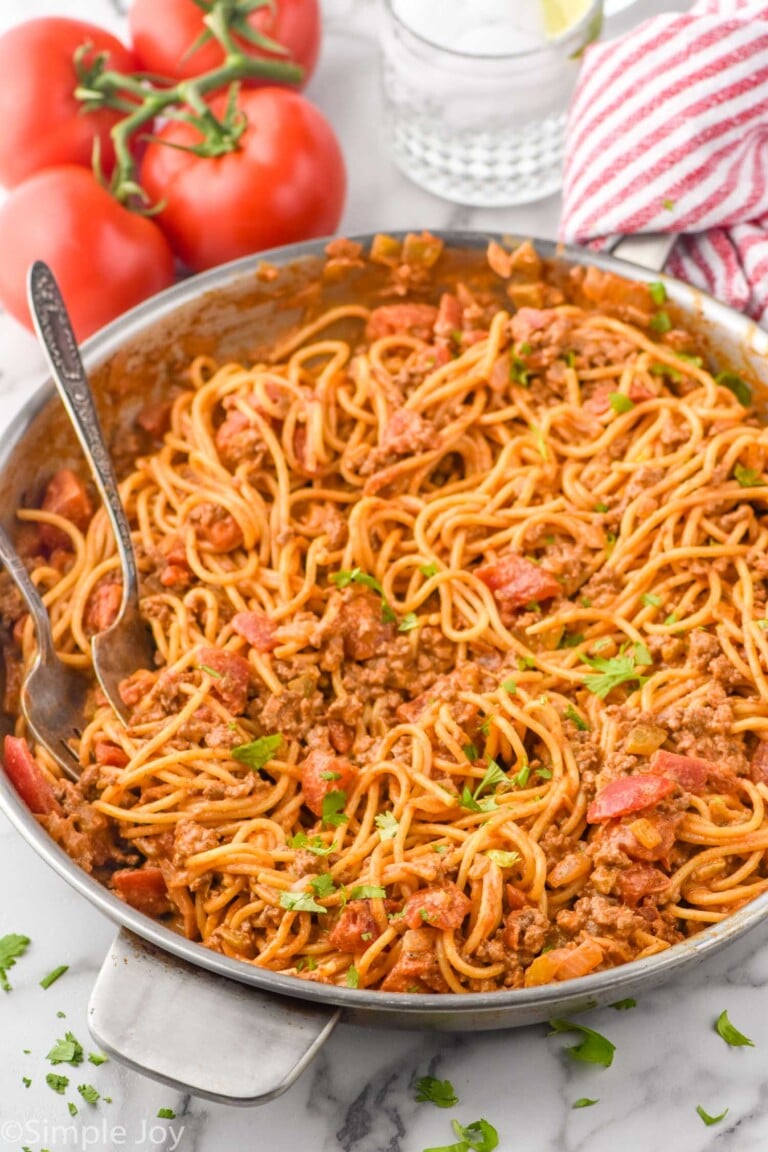 skillet of Taco Spaghetti with two forks for serving. Tomatoes and drinking glass sitting in background