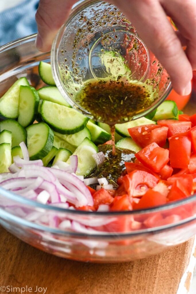 man's hand pouring small bowl of dressing into a bowl of Cucumber Tomato Salad ingredients.