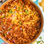 overhead of pot of Chili Mac topped with melted cheese, cilantro, and diced onion. Corn chips and fresh cilantro sitting beside