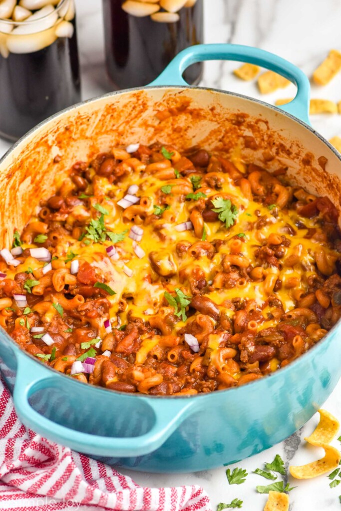 Large blue pot of chili mac topped with melted cheese, cilantro, and diced red onion