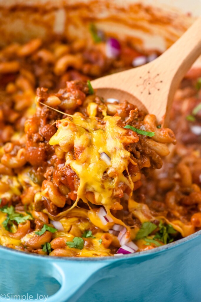 wooden spoon holding Chili Mac out of pot of Chili Mac topped with melted cheese, cilantro, and diced red onion