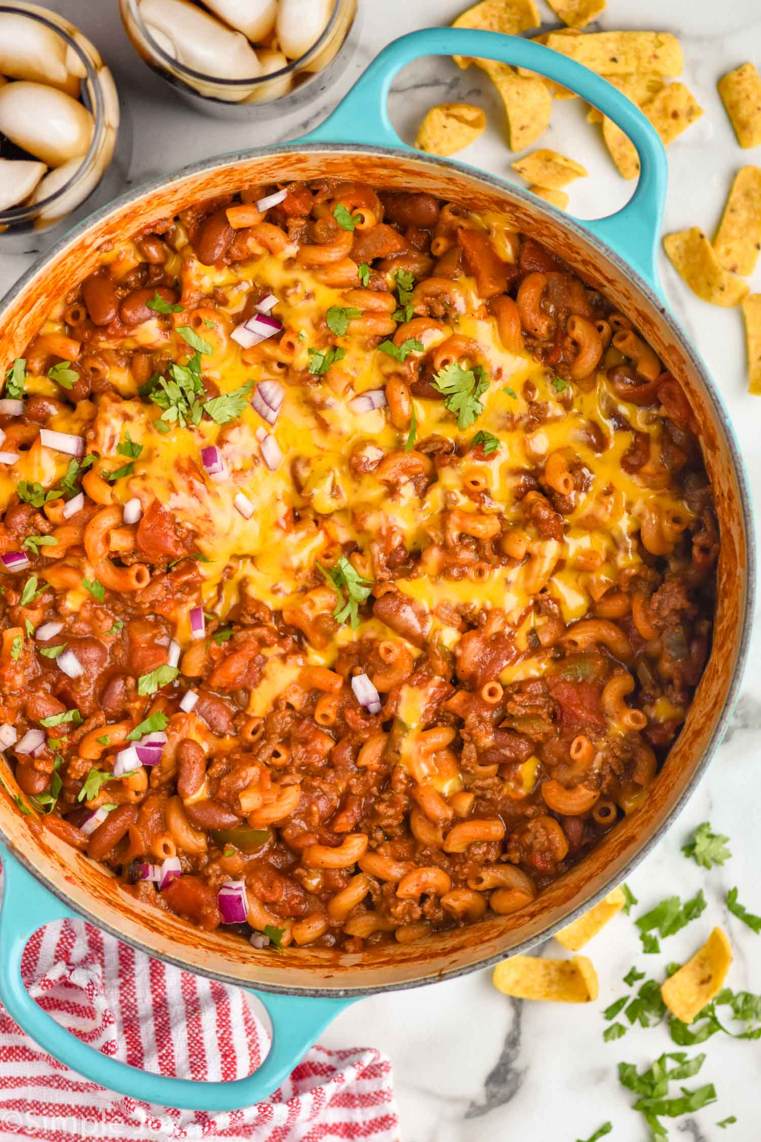 overhead of pot of Chili Mac topped with melted cheese, cilantro, and diced onion. Corn chips and fresh cilantro sitting beside