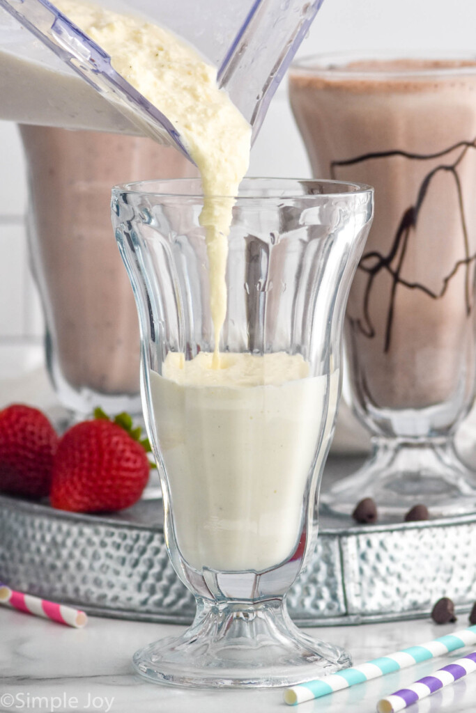 blender pouring vanilla Milkshake Recipe into a glass. chocolate milkshake, strawberry milkshake, and fresh strawberries sitting behind