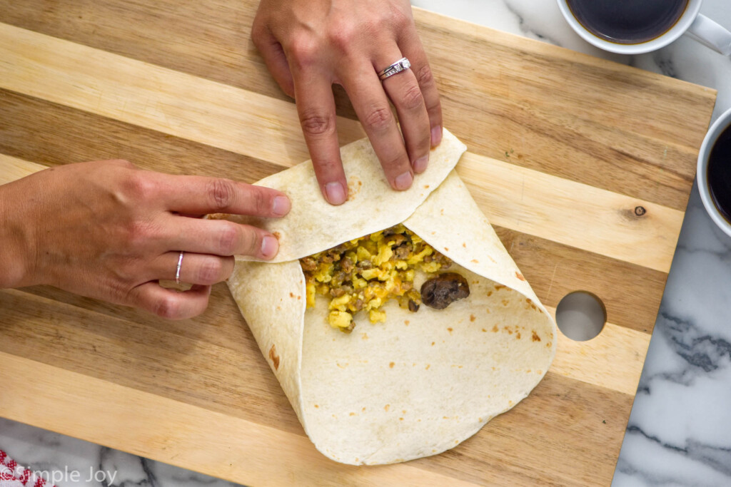 woman's hands rolling tortilla with eggs, mushrooms, and sausage inside to make a breakfast burrito