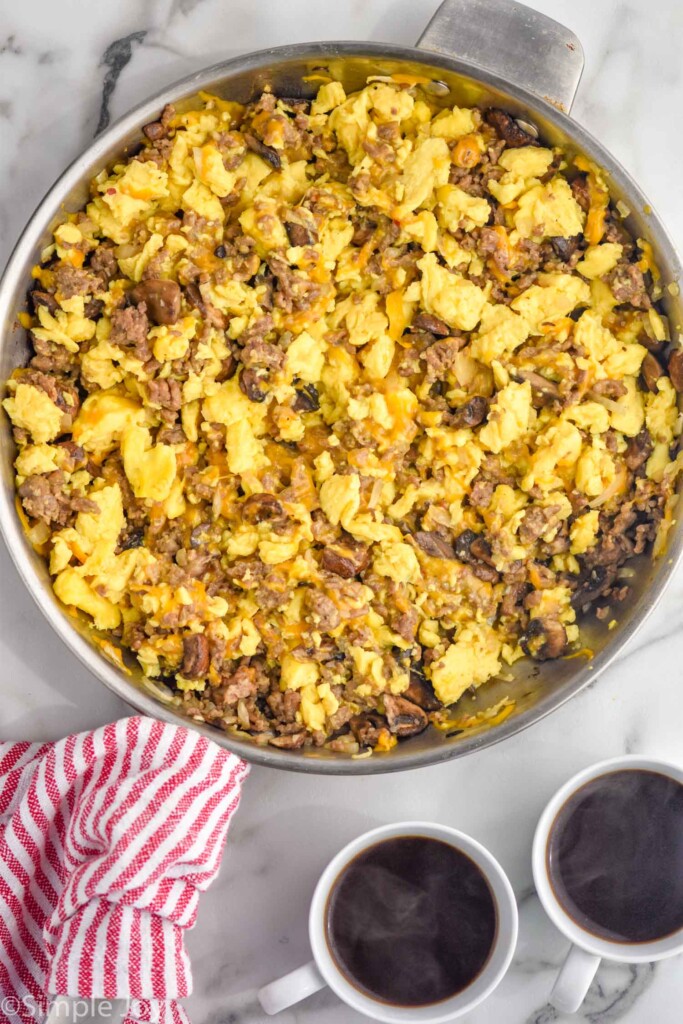 Overhead of large skillet of eggs, sausage, and cheese to make breakfast burritos. Two mugs of coffee sitting beside