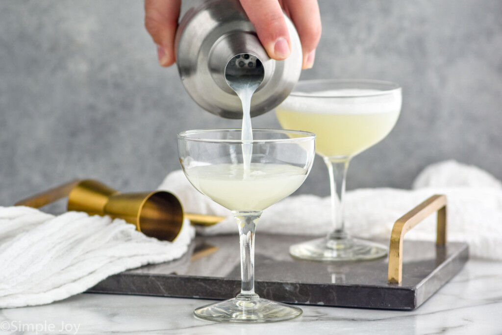 man's hand pouring cocktail shaker of pisco sour ingredients into a coupe glass. Glass of pisco sour sitting in background