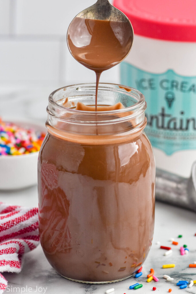 spoon pulling out of mason jar of chocolate magic shell. Sprinkles, ice cream scoop, and container of ice cream sitting in background