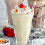 Glass of vanilla milkshake topped with whipped cream, sprinkles, and a cherry. Glasses of strawberry milkshake, chocolate milkshake, and fresh strawberries sitting in background.