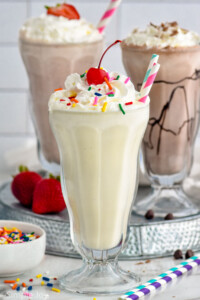 Glass of vanilla milkshake topped with whipped cream, sprinkles, and a cherry. Glasses of strawberry milkshake, chocolate milkshake, and fresh strawberries sitting in background.