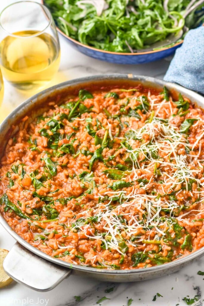 skillet of risotto with spinach and shredded parmesan cheese. Bowl of salad and glass of white wine sitting in background