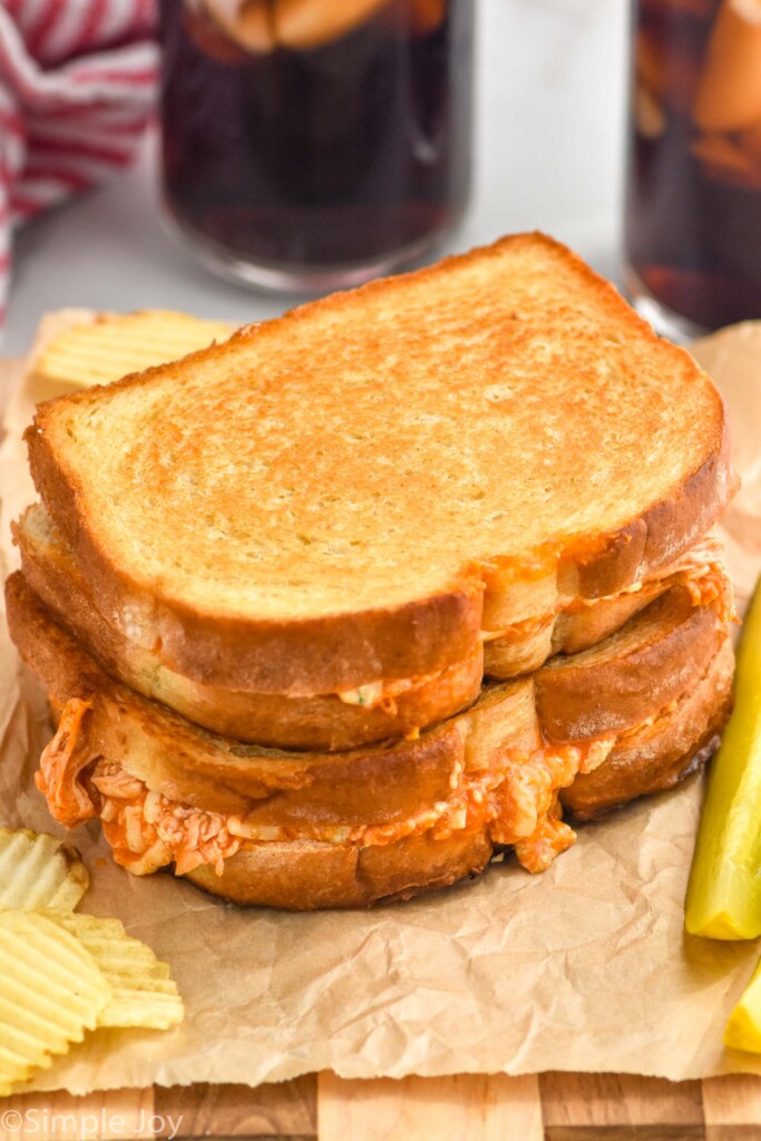 two Buffalo Chicken Sandwiches stacked on brown paper with pickle and potato chips sitting beside