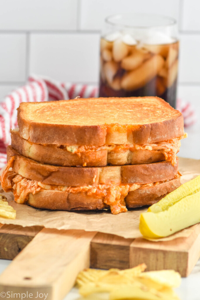 Two Buffalo Chicken Sandwiches stacked on top of each other. Pickle spear, potato chips, and drinking glass surrounding.