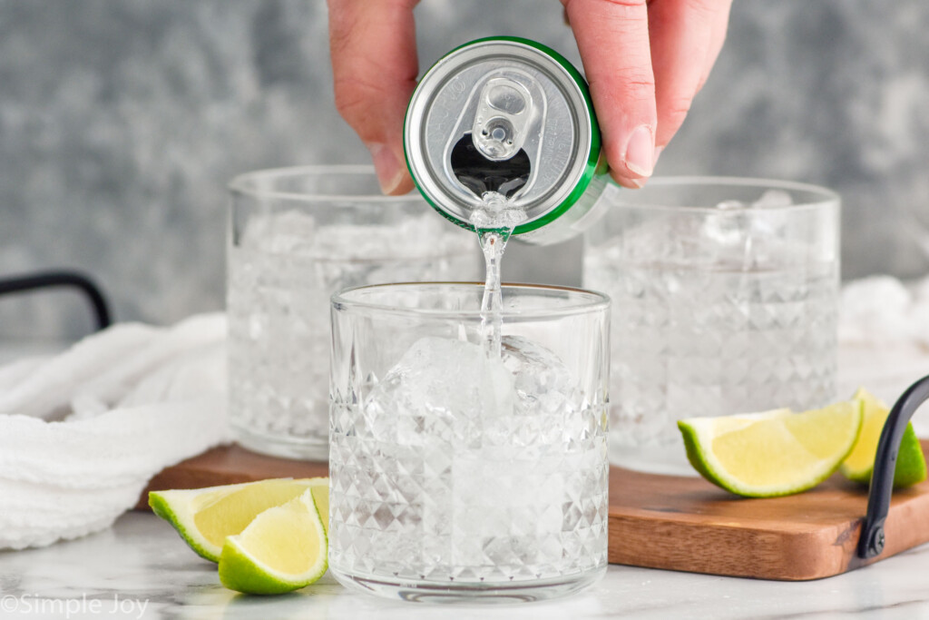 man's hand pouring can of lemon lime soda into a glass of ice and vodka press ingredients to make a vodka press. Lime wedges and two glasses of vodka press sitting behind