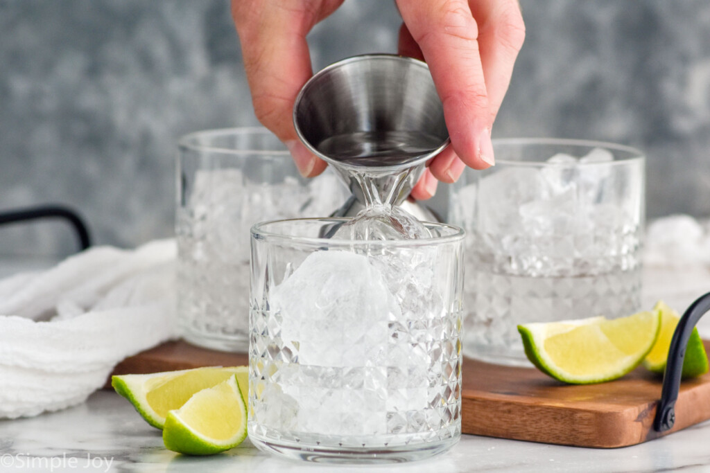 man's hand pouring vodka into a glass of ice to make a vodka press. Two glasses of ice and lime wedges sitting in background