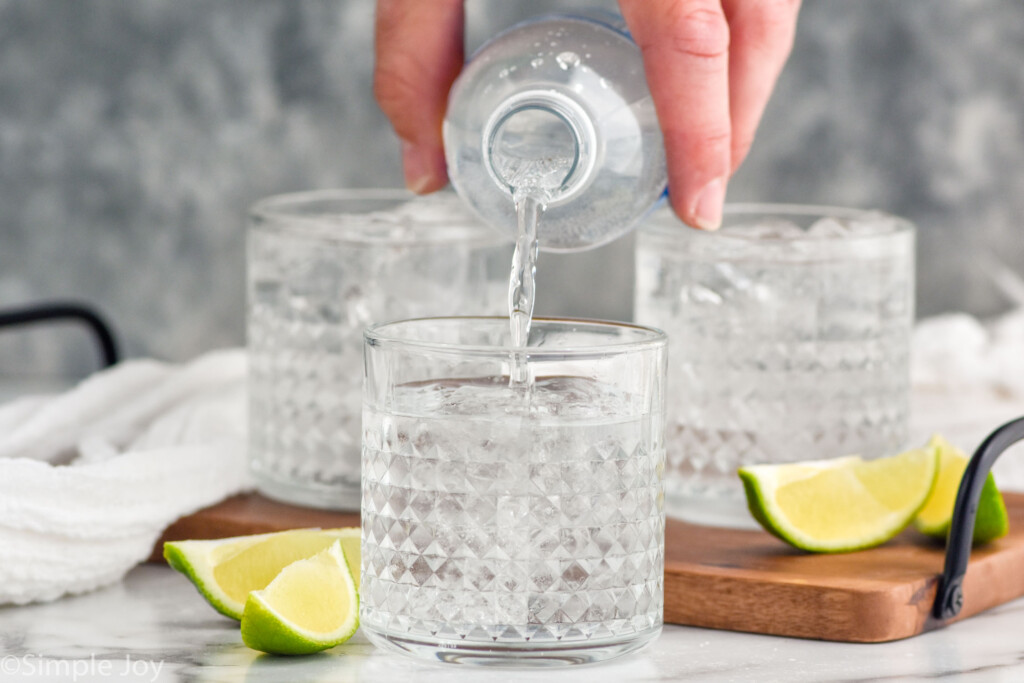 man's hand pouring bottle of club soda into a glass of vodka press ingredients. Two glasses of vodka press and lime wedges sitting behind