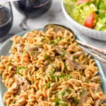 Pinterest graphic for Beef Stroganoff. Text says "the best Beef Stroganoff simplejoy.com" Image shows a serving bowl of Beef Stroganoff with two spoons for serving, bowl of lettuce salad and two glasses of red wine sitting in background.