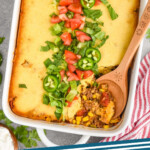 Pinterest graphic for Tamale Pie. Image shows overhead of casserole dish of Tamale Pie topped with lettuce, diced tomatoes, and jalapenos. Wooden spoon in dish for serving.